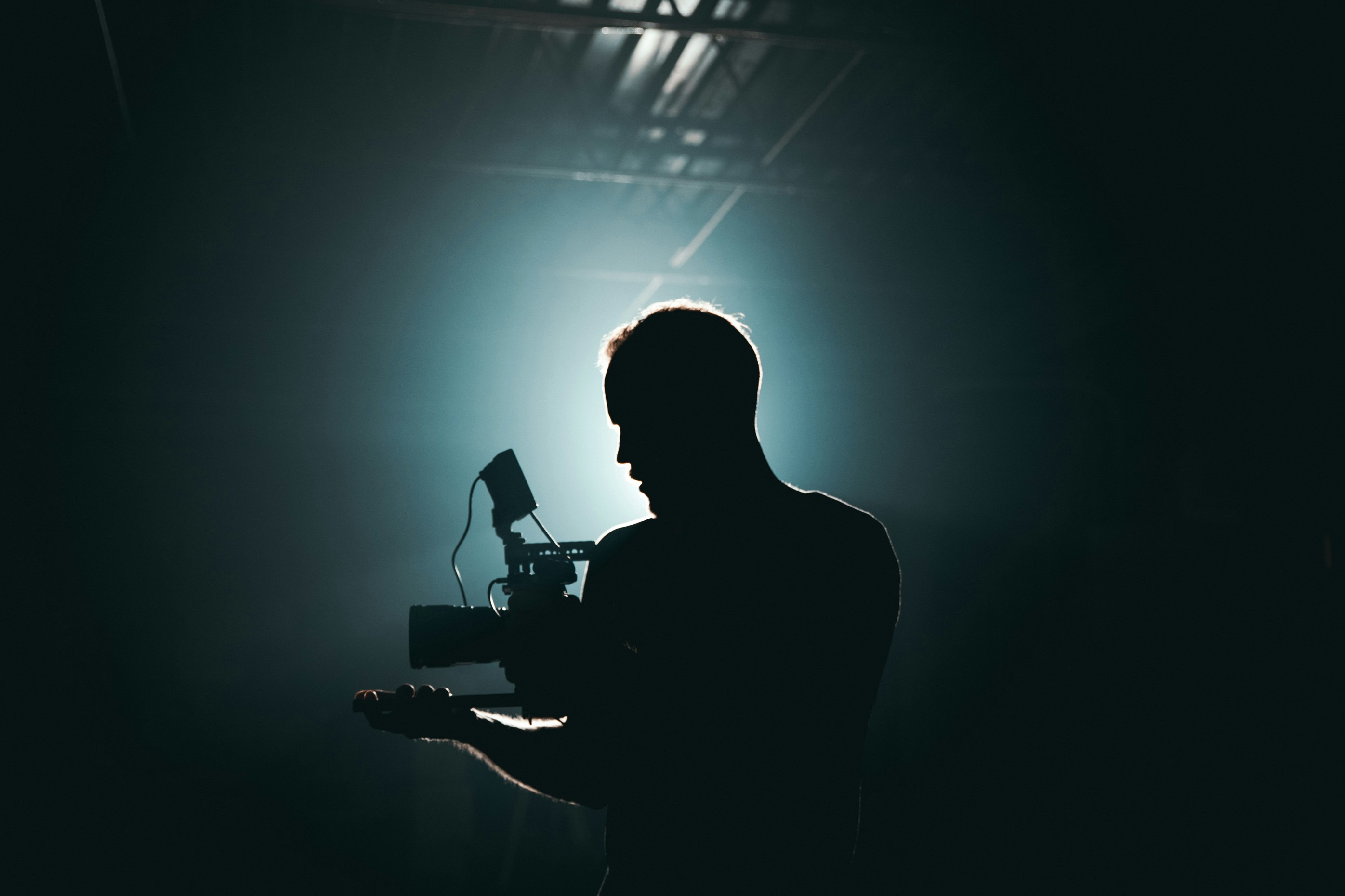 A videographer holding a camera on a dark lit set with a spotlight behind silhouetting his profile.